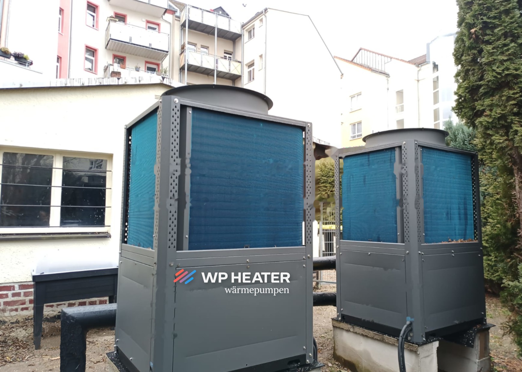 Zwei große WP-HEATER-Außeneinheiten auf Betonsockeln im Hinterhof eines Mehrfamilienhauses, mit WP-HEATER-Logo